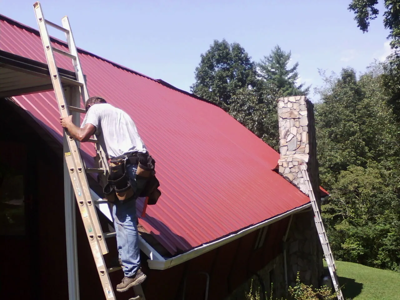 Roofer on a ladder beside a red metal roof and stone chimney — Soffit & Fascia Repair by Roofing Brothers in Niagara